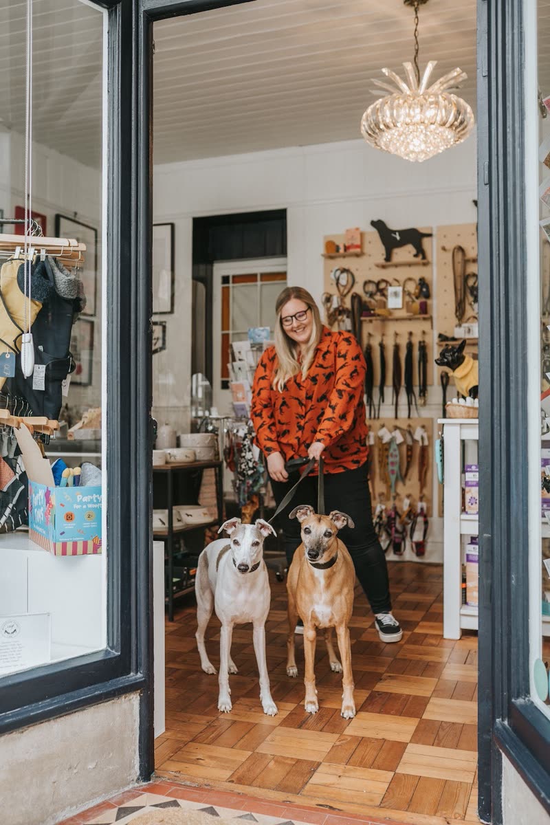 The shop owner in the doorway of the pet boutique with two whippets on leads, walls of collars and leads behind her